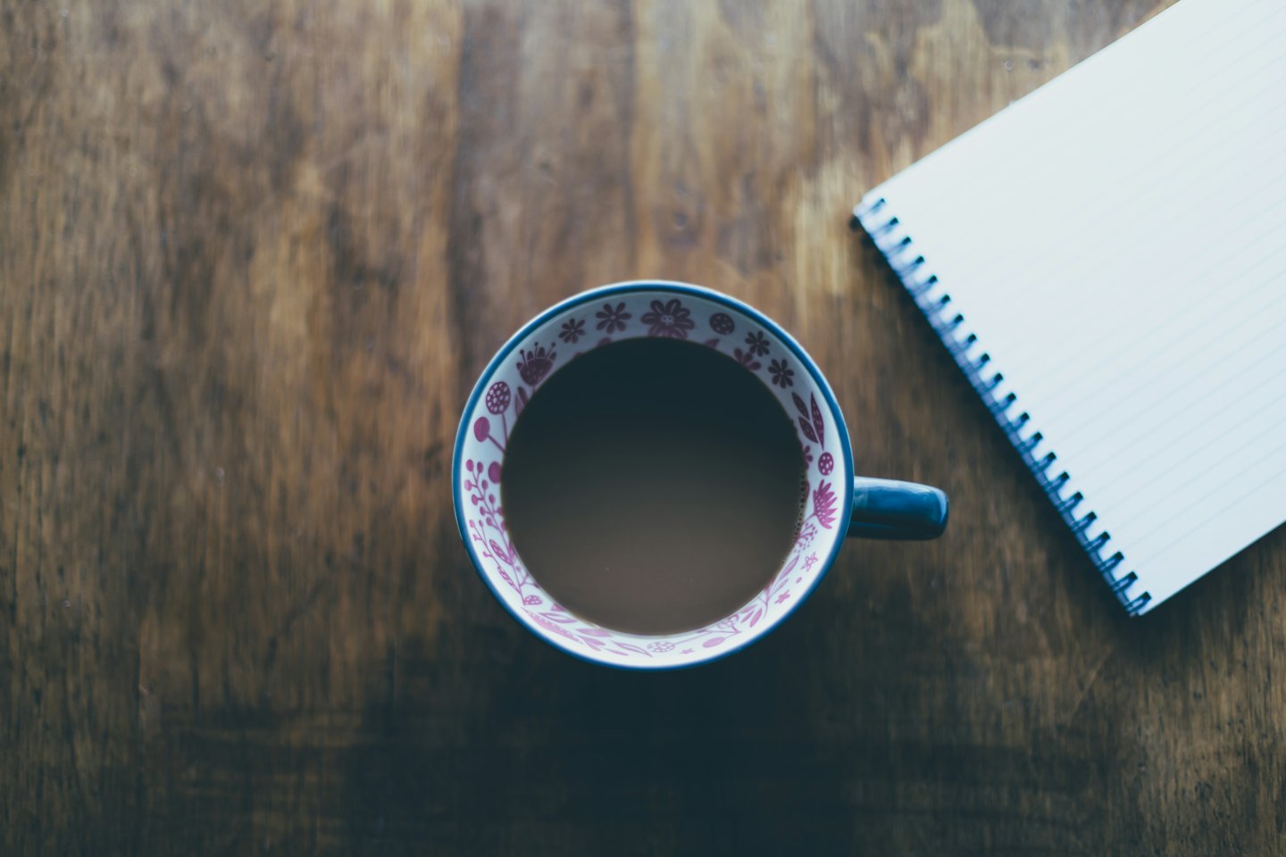 Patterned mug of coffee beside notebook on wooden desk.